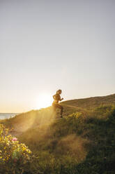 Side view of mature woman jogging on hill against sky at sunset - MASF41574