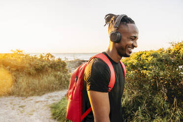 Smiling young man wearing backpack and wireless headphones against sky - MASF41583