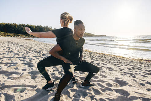 Determined man doing squats with woman on sand at beach - MASF41585