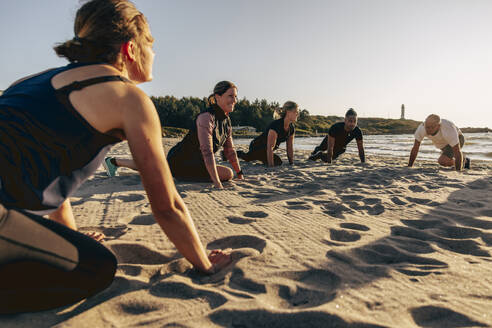 Smiling female instructor doing stretching with team on sand at beach - MASF41593