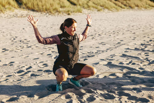 Mature female instructor explaining exercise posture while crouching on sand at beach - MASF41594