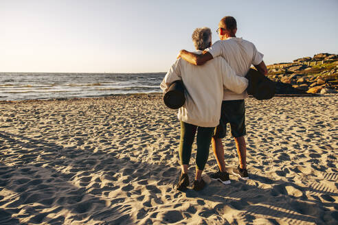 Rear view of senior couple holding exercise mats while standing on sand at beach - MASF41600
