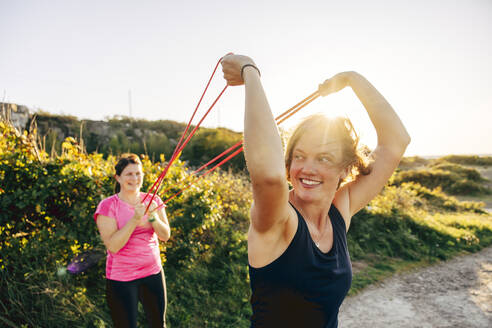 Smiling woman holding resistance band while exercising with woman at beach - MASF41608