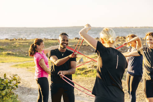 Smiling male and female teammates exercising together with resistance bands during group training session at beach - MASF41609