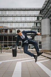 Businessman jumping mid-air while holding briefcase in front of building - MASF41611
