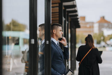 Businessman talking on smart phone while leaning on glass door at street - MASF41628