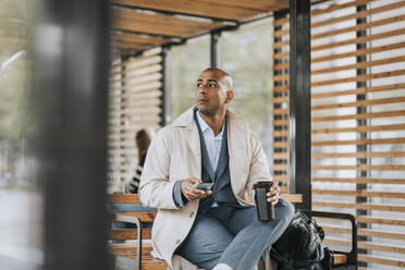 Businessman looking away holding smart phone and insulated drink container while sitting on bench at bus stop - MASF41629