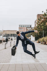 Businessman holding briefcase and jumping mid-air on footpath - MASF41637