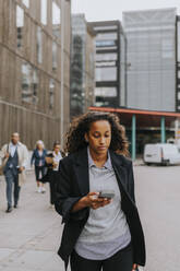 Young businesswoman using smart phone while walking on footpath in front of office building - MASF41645