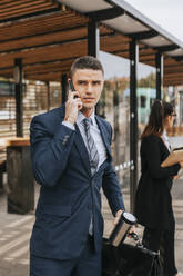 Businessman talking on smart phone while waiting at bus stop - MASF41646