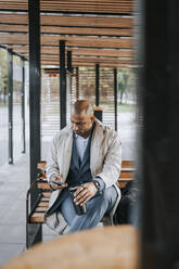Male business professional using smart phone while sitting on bench at bus stop - MASF41647