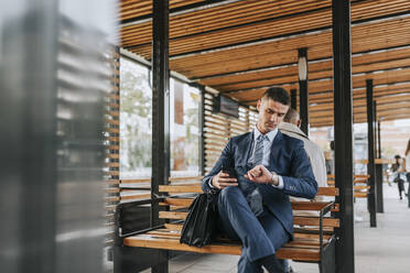 Businessman checking time while sitting on bench at bus stop - MASF41649