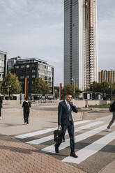 Businessman talking through smart phone while walking on zebra crossing at street in city - MASF41650