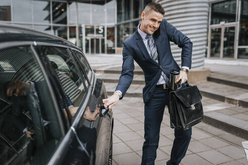 Businessman talking on smart phone while opening car door near office building - MASF41654