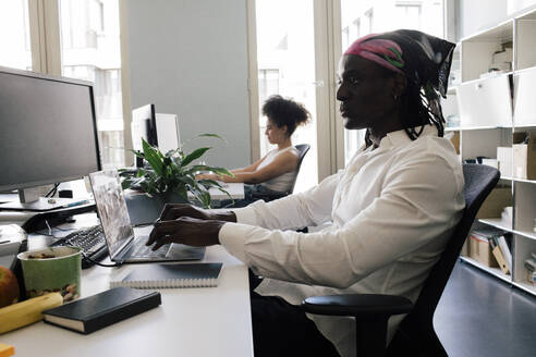 Male computer programmer using laptop while sitting on chair with colleague at office - MASF41697