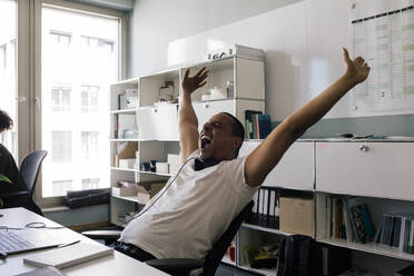 Happy male entrepreneur screaming with arms raised while sitting at office - MASF41707