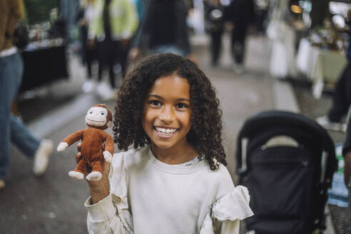Portrait of happy girl holding toy monkey at flea market - MASF41721