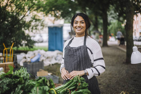 Portrait of smiling female vendor wearing apron while standing near stall at vegetable market - MASF41736