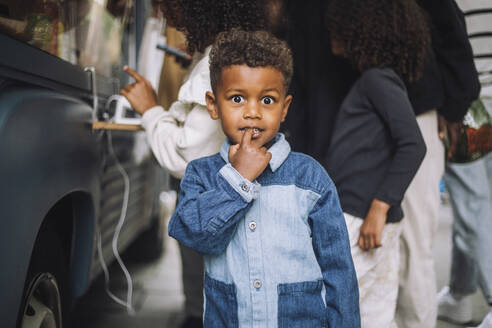 Portrait of mischievous boy standing with finger in mouth near food truck - MASF41748