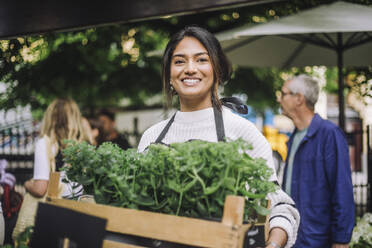 Young smiling female florist holding crate of plants at flea market - MASF41749