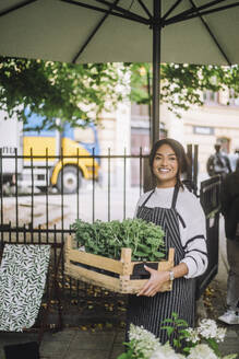 Smiling female florist holding crate of plants while standing under parasol at farmer's market - MASF41750