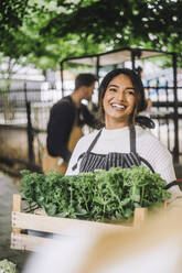 Happy female florist holding crate of plants during festival at market - MASF41751