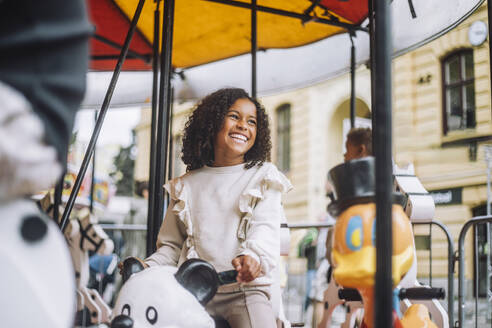 Happy girl looking away while enjoying ride on carousel during carnival at amusement park - MASF41755