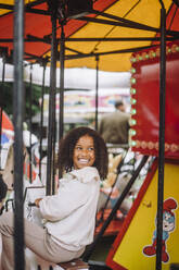 Happy girl looking over shoulder while having fun on carousel at amusement park - MASF41756