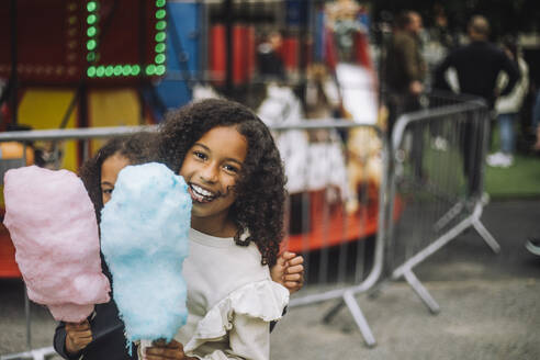 Portrait of happy girl enjoying with sister holding cotton candies at amusement park - MASF41759