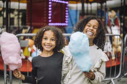 Portrait of cheerful sisters holding cotton candies while standing in front of carousel at amusement park - MASF41760