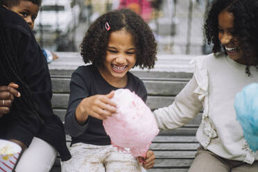 Cheerful girl having fun with siblings while holding cotton candy at amusement park - MASF41763
