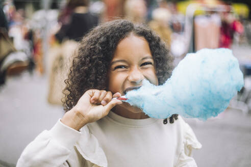 Portrait of cheerful girl eating blue cotton candy at amusement park - MASF41766