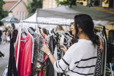 Young woman choosing dress hanging on clothes rack at flea market - MASF41771