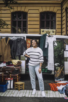Portrait of happy female owner standing with hand on hip in front of van during second hand sale at flea market - MASF41778