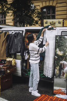 Side view full length of young female owner hanging dress during second hand sale at flea market - MASF41779