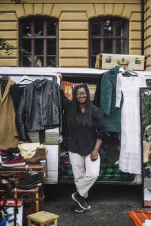 Smiling female owner standing near van while selling goods during second hand sale at flea market - MASF41782