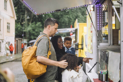 Side view of man standing with family buying snacks from food truck during carnival at amusement park - MASF41784