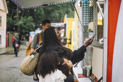 Smiling woman doing online payment near food truck while standing with family at amusement park - MASF41785