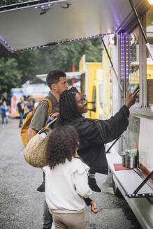 Mother doing contactless payment near food truck while standing with family at amusement park - MASF41786