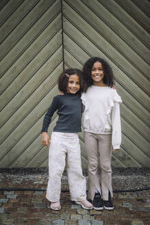 Portrait of smiling female siblings standing with arms around in front of wall - MASF41787