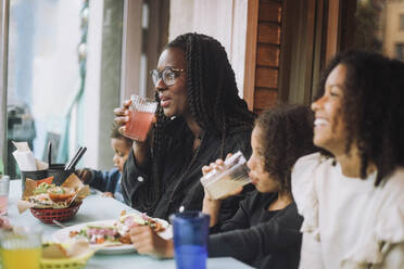 Smiling woman enjoying food and drinks with children while sitting at restaurant - MASF41791