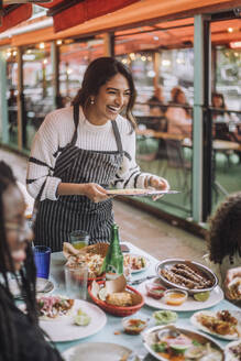 Happy waitress laughing while talking to customers near table at restaurant - MASF41813