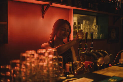 Female bartender filling beer in glass through tap while working at bar - MASF41815
