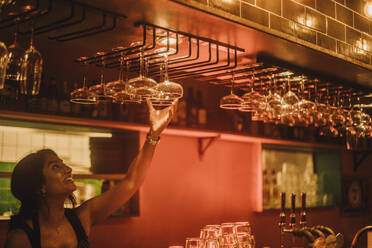 Smiling female bartender hanging cocktail glasses on rack while working at bar - MASF41816