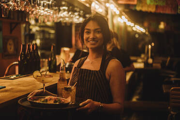 Portrait of smiling waitress holding food and drinks on serving tray while working at bar - MASF41818
