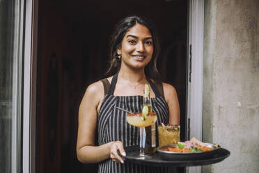 Portrait of smiling waitress holding tray of food and drinks at bar doorway - MASF41819