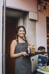 Portrait of smiling waitress wearing apron holding tray of food and drinks at bar - MASF41820