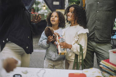 Smiling female siblings looking up while holding toys at flea market - MASF41834