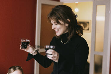 Smiling young woman holding drink glasses in apartment - MASF41850