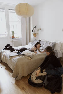 Smiling woman unpacking luggage near female friend lying down on bed in apartment - MASF41858
